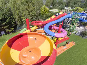 Bunte Wasserrutschen und ein Kinderbecken auf dem Campingplatz, umgeben von Rasen und Bäumen.