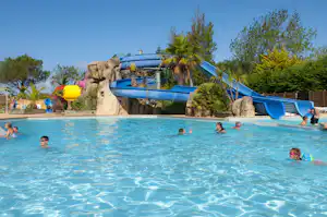 Blaues Schwimmbecken mit Kindern im Wasser und blauen Rutschen über Felsattrappe am Campingplatz.