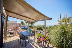 Terrasse am Campingplatz mit Tisch, Stühlen und Fahrrad, daneben ein Auto und Blick ins Grüne und auf die Berge.