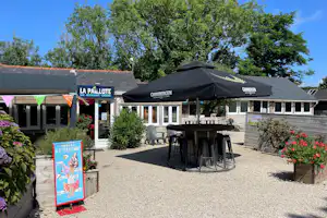 Terrasse am Campingplatz mit zwei schwarzen Marktschirmen, Tisch und Stühlen vor einem Gebäude und Hecken.