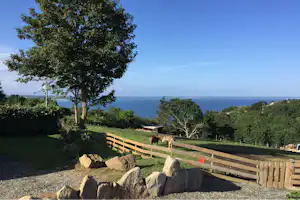 Blick vom Campingplatz über das Meer, mit Holzzaun, Steinen und Bäumen im Vordergrund.