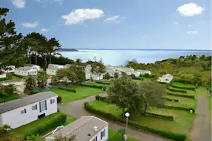 Blick auf einen Campingplatz mit weißen Mobilheimen, gepflegten Wegen und Rasenflächen sowie Meerblick und Bäumen am Horizont.