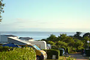 Wohnmobil auf einem Stellplatz mit Hecken und Bäumen, im Hintergrund das Meer und ein Wohnwagen im Campingbereich.