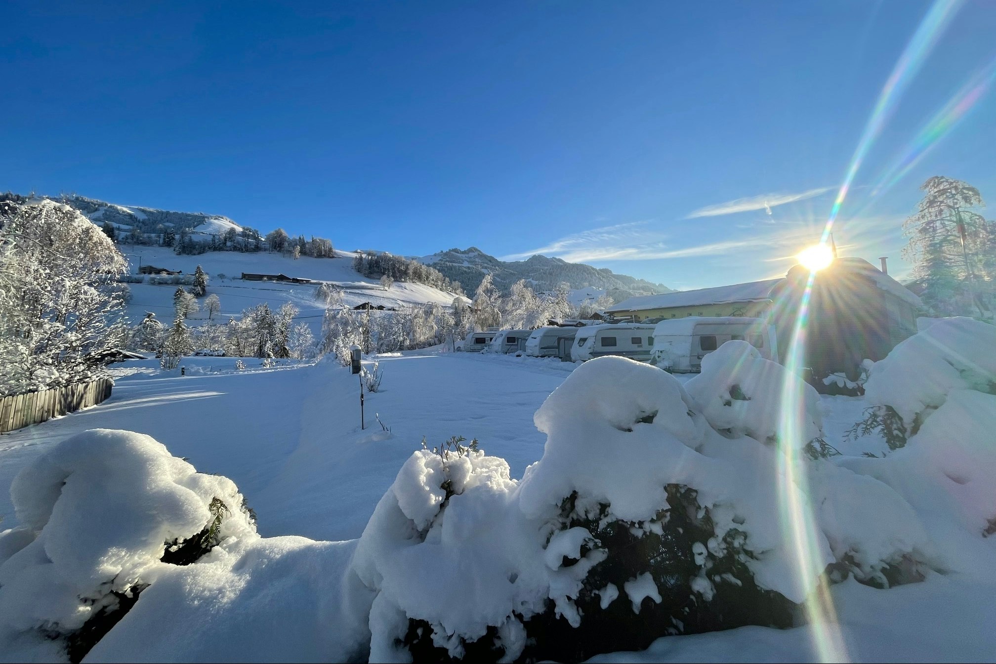 Verschneite Landschaft mit verschneiten Büschen und einem weißen Gebäude im Hintergrund, Berge und klarer Himmel bei tiefstehender Sonne.