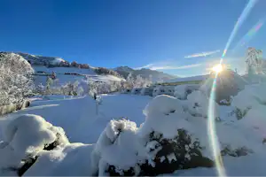 Verschneite Landschaft mit verschneiten Büschen und einem weißen Gebäude im Hintergrund, Berge und klarer Himmel bei tiefstehender Sonne.