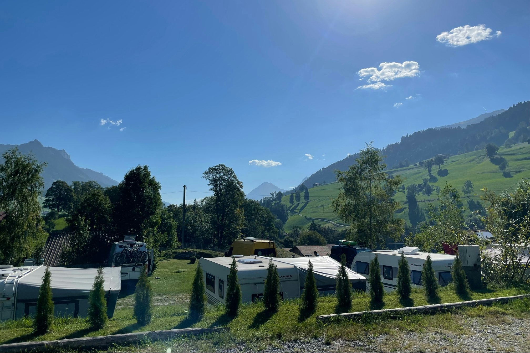 Reihen von Wohnwagen und Stellplätzen auf einem Campingplatz, mit Hecken, Bäumen und Blick auf grüne Berge unter blauem Himmel.