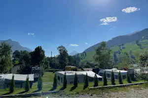 Reihen von Wohnwagen und Stellplätzen auf einem Campingplatz, mit Hecken, Bäumen und Blick auf grüne Berge unter blauem Himmel.
