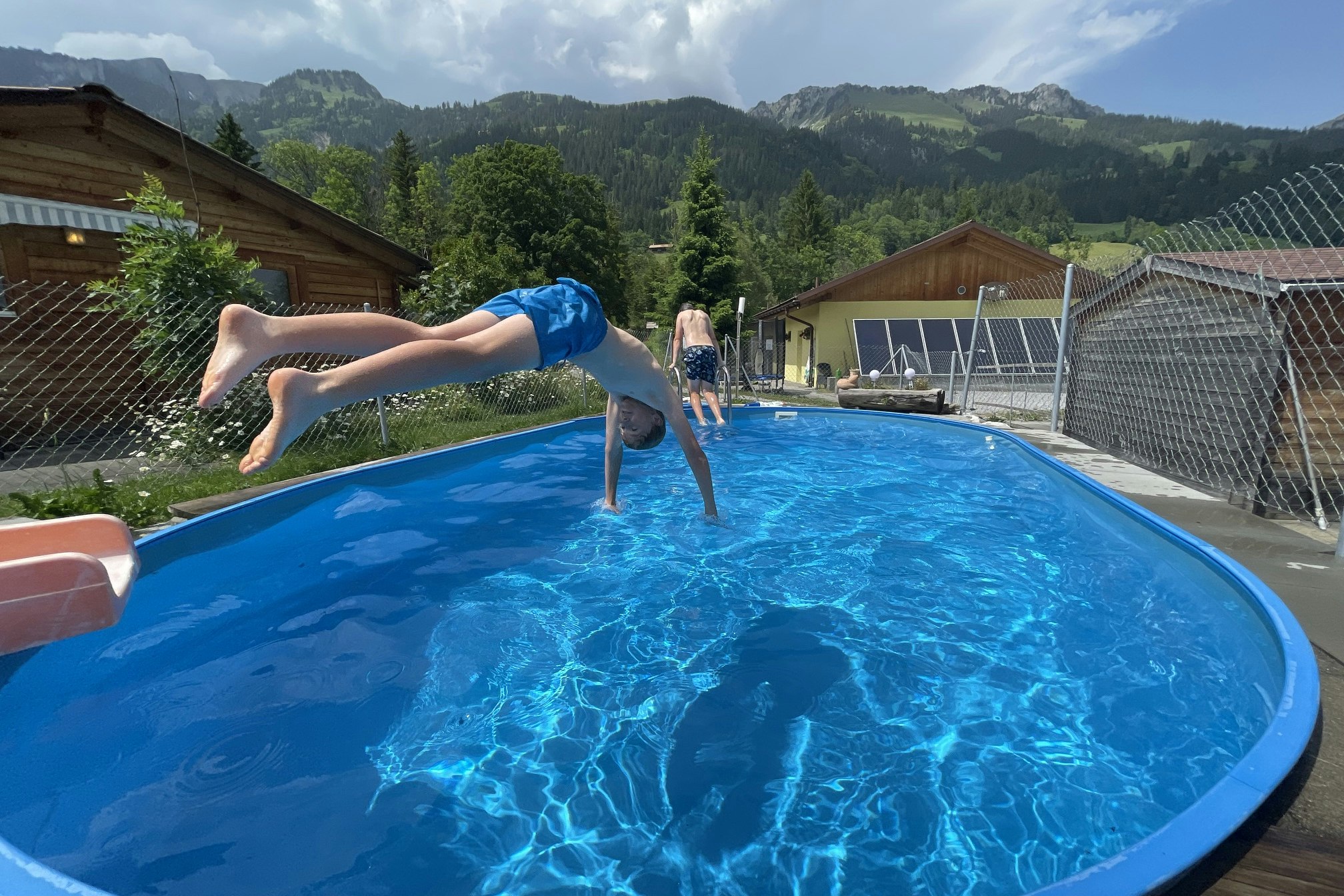 Kinder springen im blauen Rundpool, im Hintergrund Zäune, Gebäude und Berge mit grünen Wäldern.