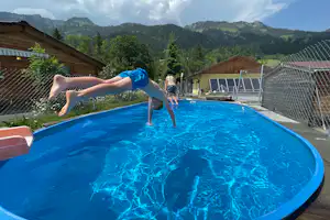 Kinder springen im blauen Rundpool, im Hintergrund Zäune, Gebäude und Berge mit grünen Wäldern.