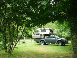 Grünes Wohnmobil auf einem Campingplatz zwischen Bäumen, mit Tisch und Stühlen im Gras.