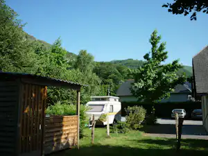 Wohnwagen im Campinggarten mit Rasen, Holzsichtschutz, Grill und Sitzbank, umgeben von Bäumen und Hügeln im Hintergrund.