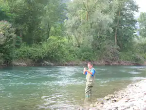 Ein Mann steht im flachen Wasser eines Flusses, umgeben von Bäumen und grünem Ufer, mit Steinen am Rand.