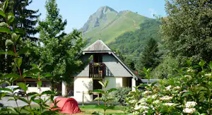 Campinghaus mit Holzveranda, umgeben von Bäumen und Blumenbeeten, im Hintergrund grüne Berge.