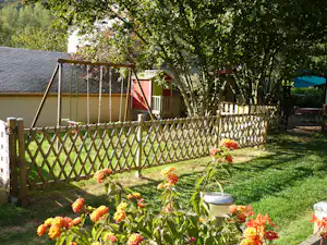 Blumenbeet im Campinggarten vor einem Holzlattenzaun, dahinter ein Wohnwagen mit Veranda und eine Schaukel unter Bäumen.