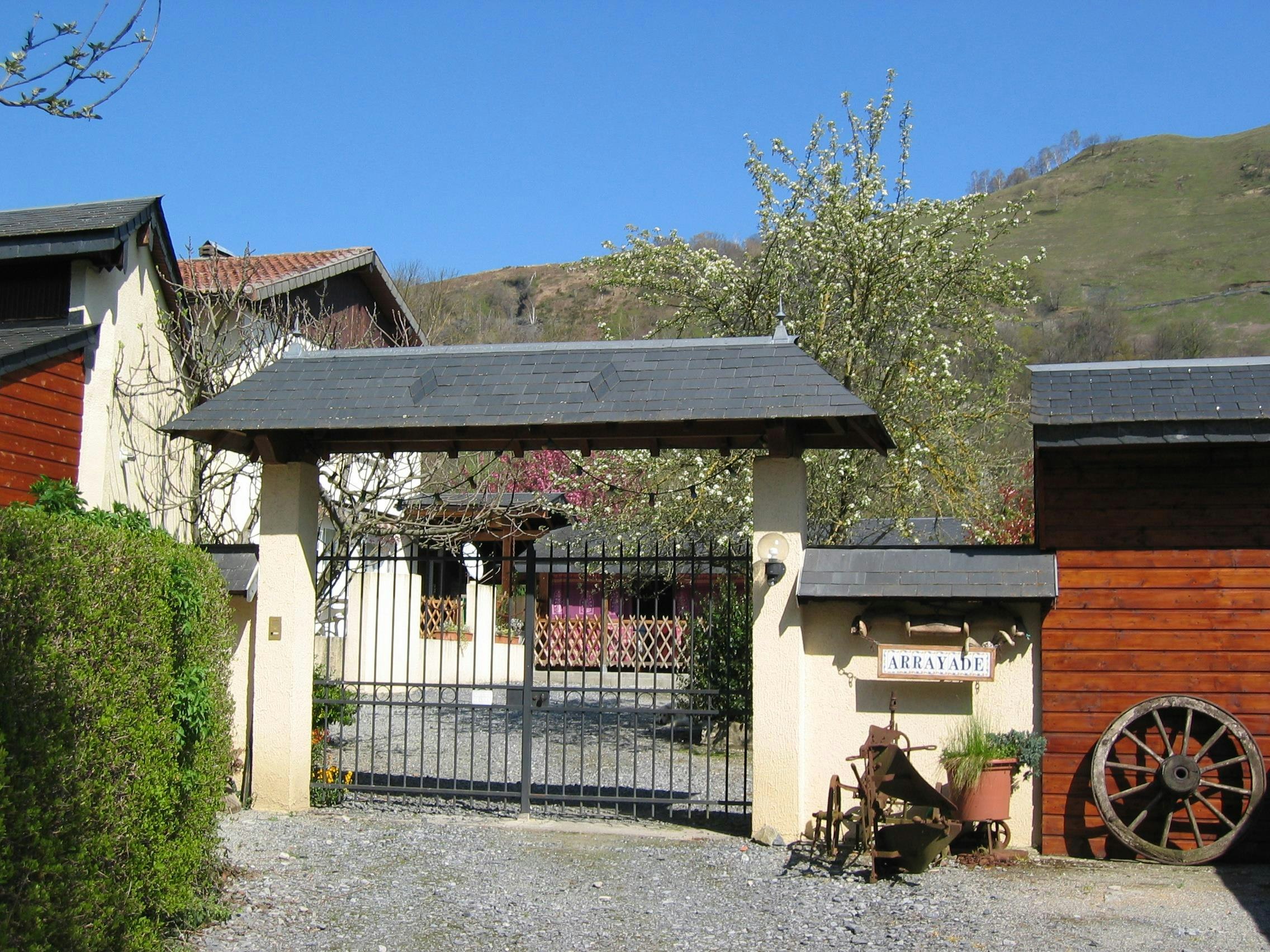 Camping-Eingang mit Tor, Ziegelwand und Holzschuppen, Kiesweg und Pflanzen vor dem Gebäude, im Hintergrund grüne Hügel.