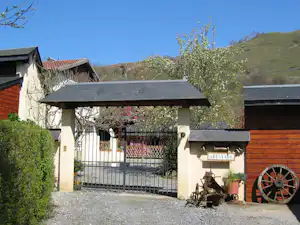 Camping-Eingang mit Tor, Ziegelwand und Holzschuppen, Kiesweg und Pflanzen vor dem Gebäude, im Hintergrund grüne Hügel.