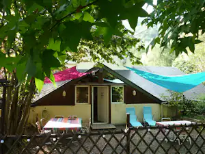 Kleines Campinghaus mit Holzveranda, Tisch und Stühlen, zwei Liegestühlen und Sonnenschirm im Schatten von Bäumen.
