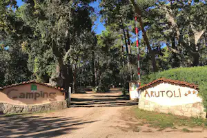 Eingang eines Campingplatzes mit Schild „ARUTOLI“, Steinmauer und Weg durch Bäume im Hintergrund.