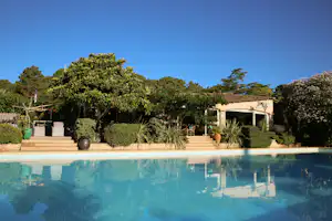 Schwimmbecken mit Spiegelung, im Hintergrund Bäume und ein Poolbereich mit Sonnenschirm und Liegen.