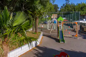 Spielplatz auf dem Campingplatz mit Rutsche, Klettergerüst und eingezäuntem Bereich, umgeben von Palmen und Bäumen.