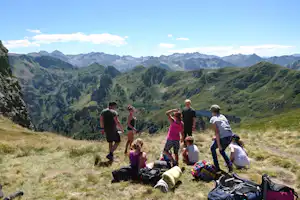 Wandergruppe mit Rucksäcken auf einer Wiese vor einem Bergtal mit See und grünen Hängen.
