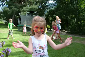 Kind mit geschminktem Gesicht auf einer grünen Wiese, im Hintergrund Kinder und ein Trampolin im Campingbereich.