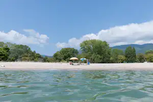 Blick auf den See mit Strand, im Hintergrund ein Sonnenschirm und ein kleines Zelt am Ufer.