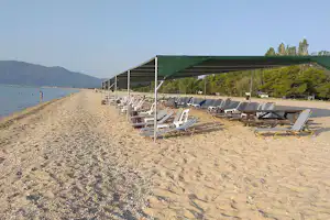Reihe Strandliegen unter Sonnensegeln am Campingplatz, Sandstrand mit Blick auf das Wasser und Berge in der Ferne.