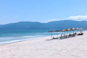 Weißer Sandstrand mit Sonnenschirmen und Liegen, ruhige Wellen und Berge im Hintergrund.