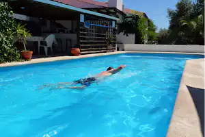 Blauer Camping-Pool mit schwimmender Person, daneben Liegen und Tische an der Terrasse vor dem Haus.