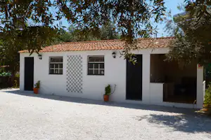 Weißes Campinghaus mit Terrasse, zwei Fenstern und Schattenbereich, davor Kiesfläche und Töpfe an der Wand.