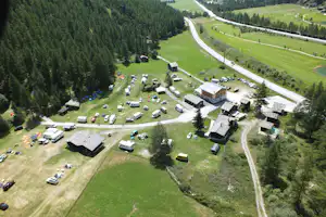 Luftaufnahme eines Campingplatzes mit Wohngebäuden, vielen Stellplätzen, Zelten und Autos auf einer Wiese am Waldrand.