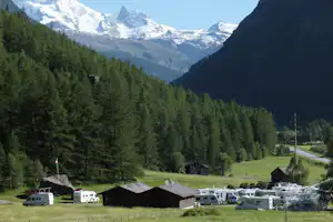 Campingplatz im Alpen-Tal: Wohnmobile und Gebäude am Rand, dahinter Wald und hohe schneebedeckte Berge.