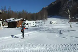 Winterlicher Campingplatz mit Wohnmobilen, verschneiten Wegen und einem Skifahrer vor einem Holzgebäude im Tal.