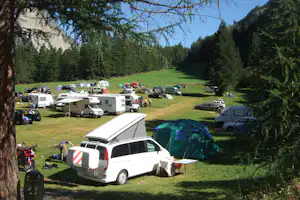 Campingplatz mit Wohnmobilen, Autos und Zelten auf einer grünen Wiese, umgeben von Bäumen und Bergen im Hintergrund.