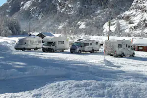 Mehrere Wohnmobile auf einem verschneiten Campingplatz vor einer Felswand und einem kleinen Gebäude.