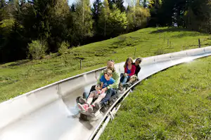 Familie auf einer Wasserrutsche auf einer grünen Wiese, umgeben von Bäumen und einem Hang mit Zäunen.