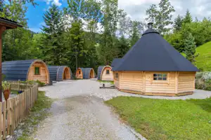Holzblockhütte mit Schindeldach und Eingang, daneben mehrere Zeltbungalows auf Kiesweg im grünen Campingbereich.
