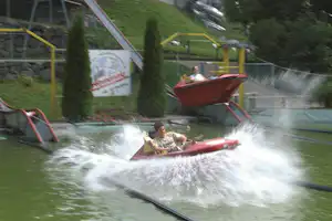Mann im roten Schlauchboot auf einer Wasserbahn, Spritzwasser und Rutsche im Campingbereich, im Hintergrund Bäume und Zäune.