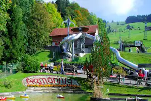 Wasserpark am Campingplatz mit Rutsche, Gebäude und Blumenbeet am Teich, umgeben von grünen Bäumen und Wiesen.