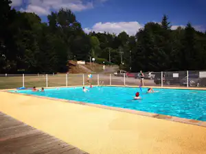 Blaues Camping-Schwimmbecken mit Zaun, Liegen und mehreren Personen im Wasser, dahinter Bäume und ein Weg.