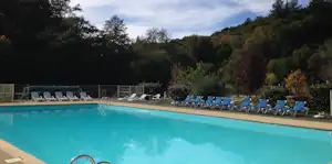 Schwimmbad mit türkisfarbenem Wasser und Liegen am Rand, umgeben von Bäumen und einem Zaun.