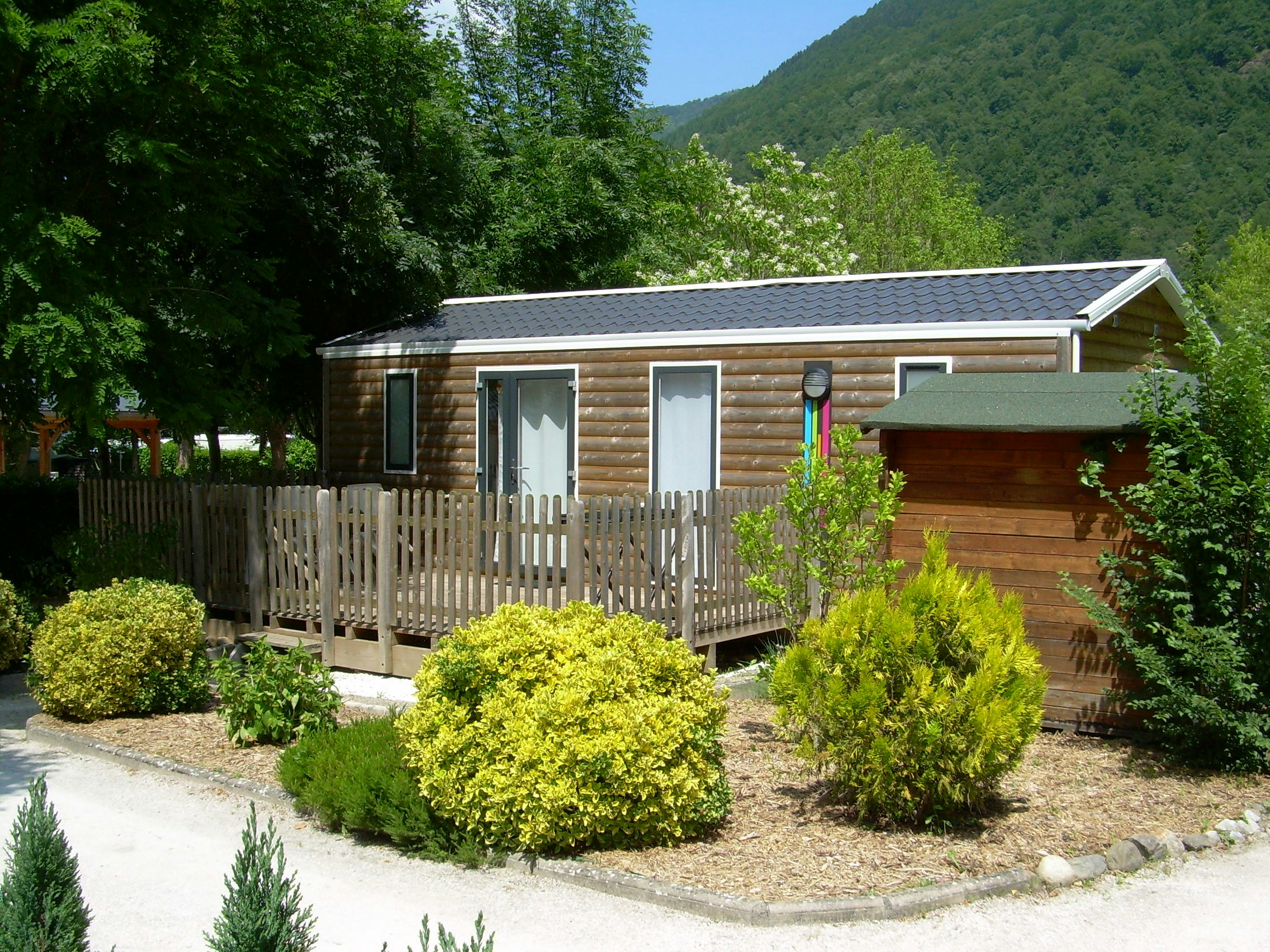 Holzbungalow mit Terrasse und Stufen, umgeben von Sträuchern und Wegen, im Hintergrund grüne Berge und Bäume.