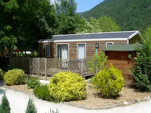 Holzbungalow mit Terrasse und Stufen, umgeben von Sträuchern und Wegen, im Hintergrund grüne Berge und Bäume.