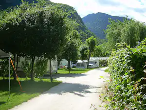 Campingplatz-Allee mit Bäumen, Kiesweg und Wohnmobilen, im Hintergrund grüne Berge und bewölkter Himmel.