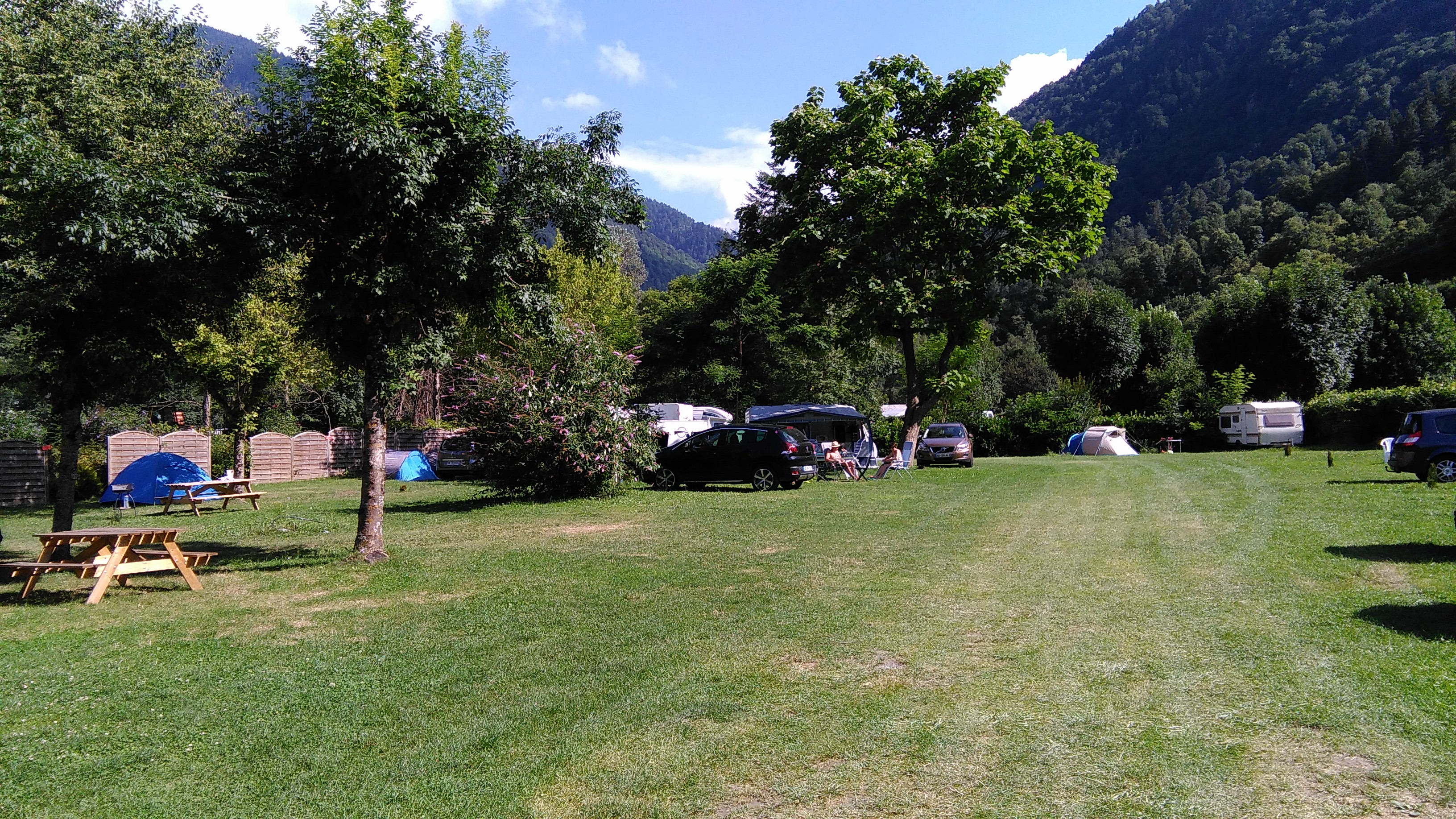 Campingwiese mit Wohnwagen und mehreren Autos, Bäume und Berge im Hintergrund.