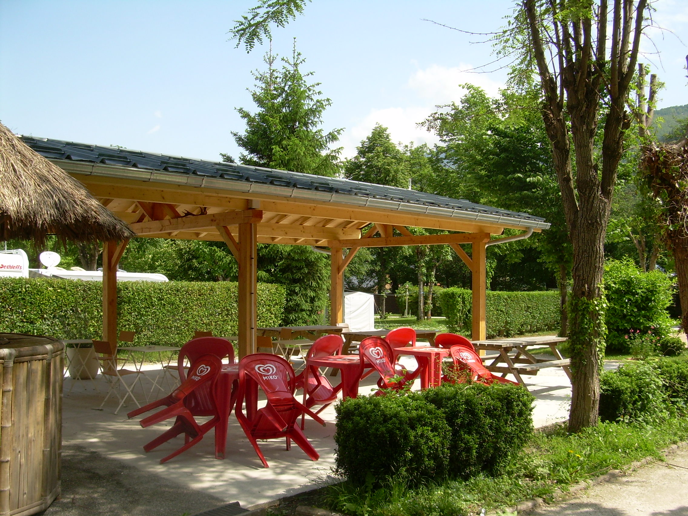 Überdachte Terrasse mit Holzdach, roten Kunststoffstühlen und Tisch, Hecke und Bäumen am Campingplatz