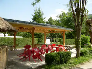 Überdachte Terrasse mit Holzdach, roten Kunststoffstühlen und Tisch, Hecke und Bäumen am Campingplatz
