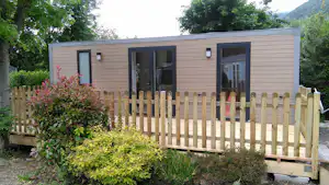 Bungalow mit Holzsichtschutzzaun und Terrasse, umgeben von Büschen und Bäumen, im Hintergrund ein bewaldeter Hügel.