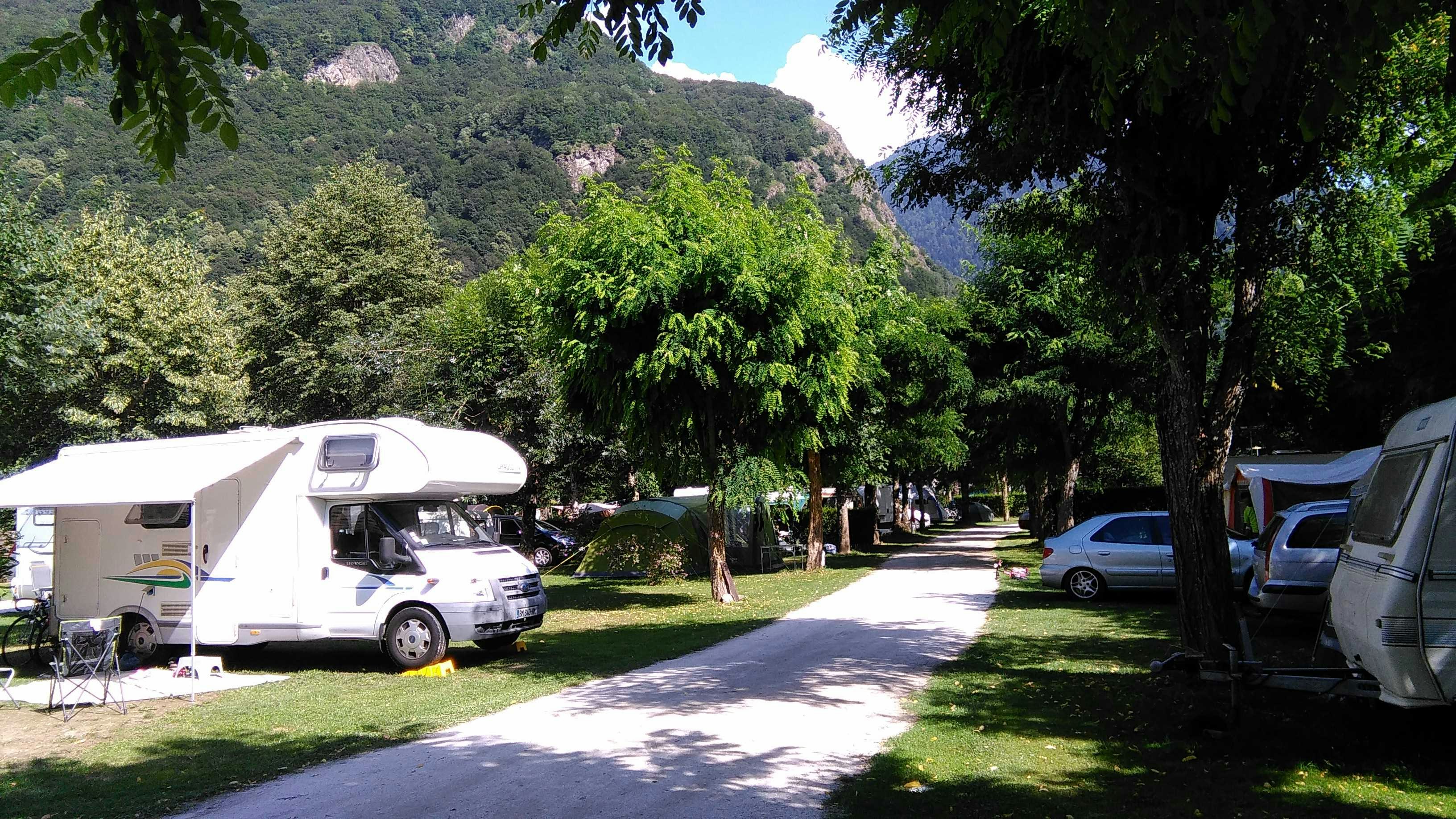Wohnwagen und Campingfahrzeuge auf einem Stellplatz im Grünen, mit Bäumen und einem Weg zwischen den Fahrzeugen.